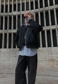 Jeune personne portant une veste noire, une chemise blanche, une cravate noire et des lunettes de soleil, ajustant ses lunettes de soleil, debout devant un mur de ventilation en pierre élevé.