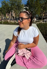 Haut blanc à manches courtes, jupe texturée rose, baskets noires et un sac à main rose clair avec un nœud. Grandes lunettes de soleil noires. Cadre extérieur.