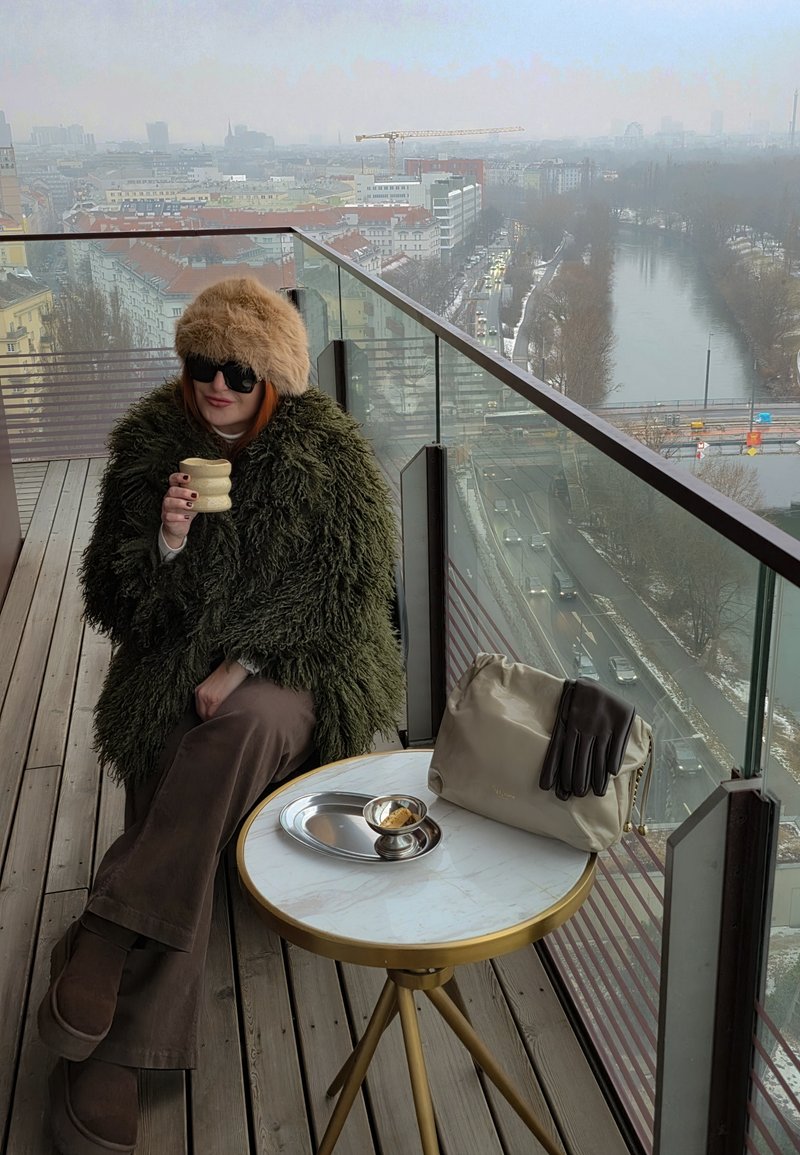 Άτομο με γούνινο παλτό και καπέλο κάθεται σε μπαλκόνι κρατώντας μια κούπα, με μια μπεζ τσάντα και γάντια πάνω σε ένα μικρό στρογγυλό τραπέζι δίπλα του.