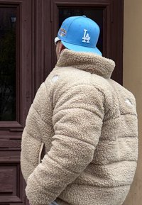 Μπεζ φλις τζάκετ με ψηλό γιακά και υφή, συνδυασμένο με ελαφρύ μπλε καπέλο μπέιζμπολ με λευκό λογότυπο "LA".