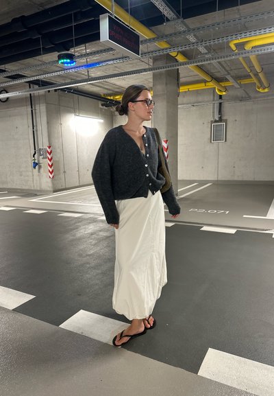 Cárdigan gris de punto con grandes botones, combinado con una falda maxi blanca y chanclas negras, en un garaje de concreto.
