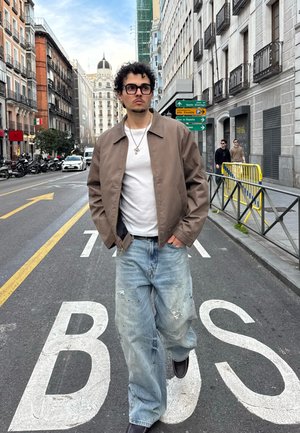 Joven con gafas, chaqueta marrón, camisa blanca y vaqueros holgados caminando por una calle de la ciudad señalizada como "BUS", con edificios y vehículos cerca.