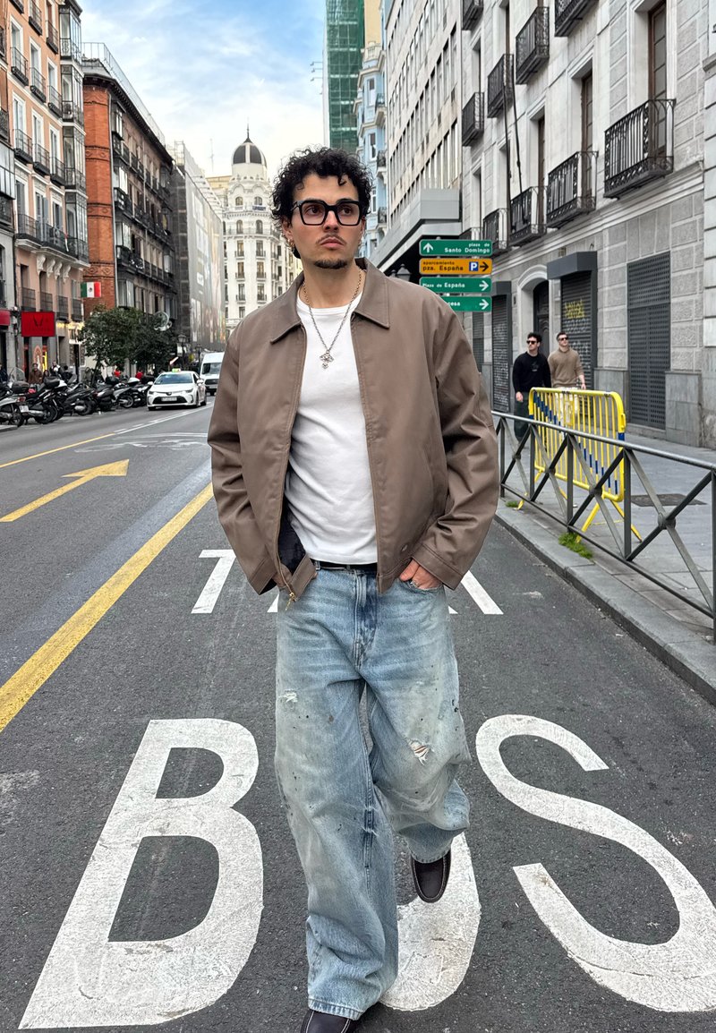 Jeune homme portant des lunettes, veste marron, chemise blanche et jean ample, marchant dans une rue de la ville marquée "BUS" avec des bâtiments et des véhicules à proximité.