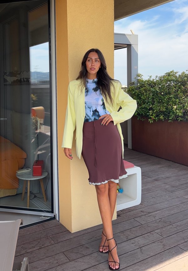 Femme aux longs cheveux foncés se tenant sur un balcon en bois, vêtue d'un blazer jaune, d'un t-shirt tie-dye, d'une jupe marron et de sandales noires à brides.