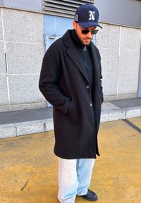 Μαύρο μάλλινο παλτό με ραμμένο γιακά, συνδυασμένο με μαύρο τζέρσεϋ, ανοιχτό μπλε φαρδιά τζιν, μαύρα παπούτσια και μπλε καπελάκι του μπέιζμπολ.