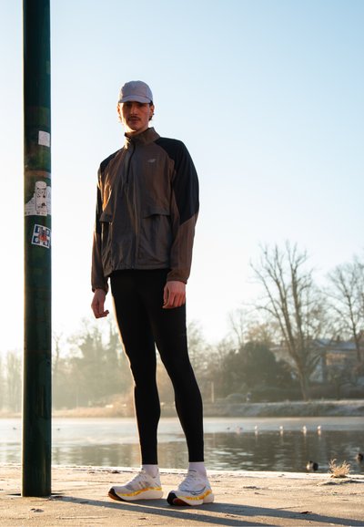 Joven con gorra, chaqueta y pantalones ajustados de pie junto a un lago al amanecer, con árboles y pájaros de fondo.
