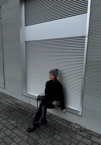 Άτομο που φοράει ένα μαύρο πουλόβερ, γκρίζο καπέλο και μαύρες μπότες μέχρι το γόνατο, καθισμένο ενάντια σε έναν υφασμάτινο ασημί τοίχο με γεωμετρικό σχέδιο.