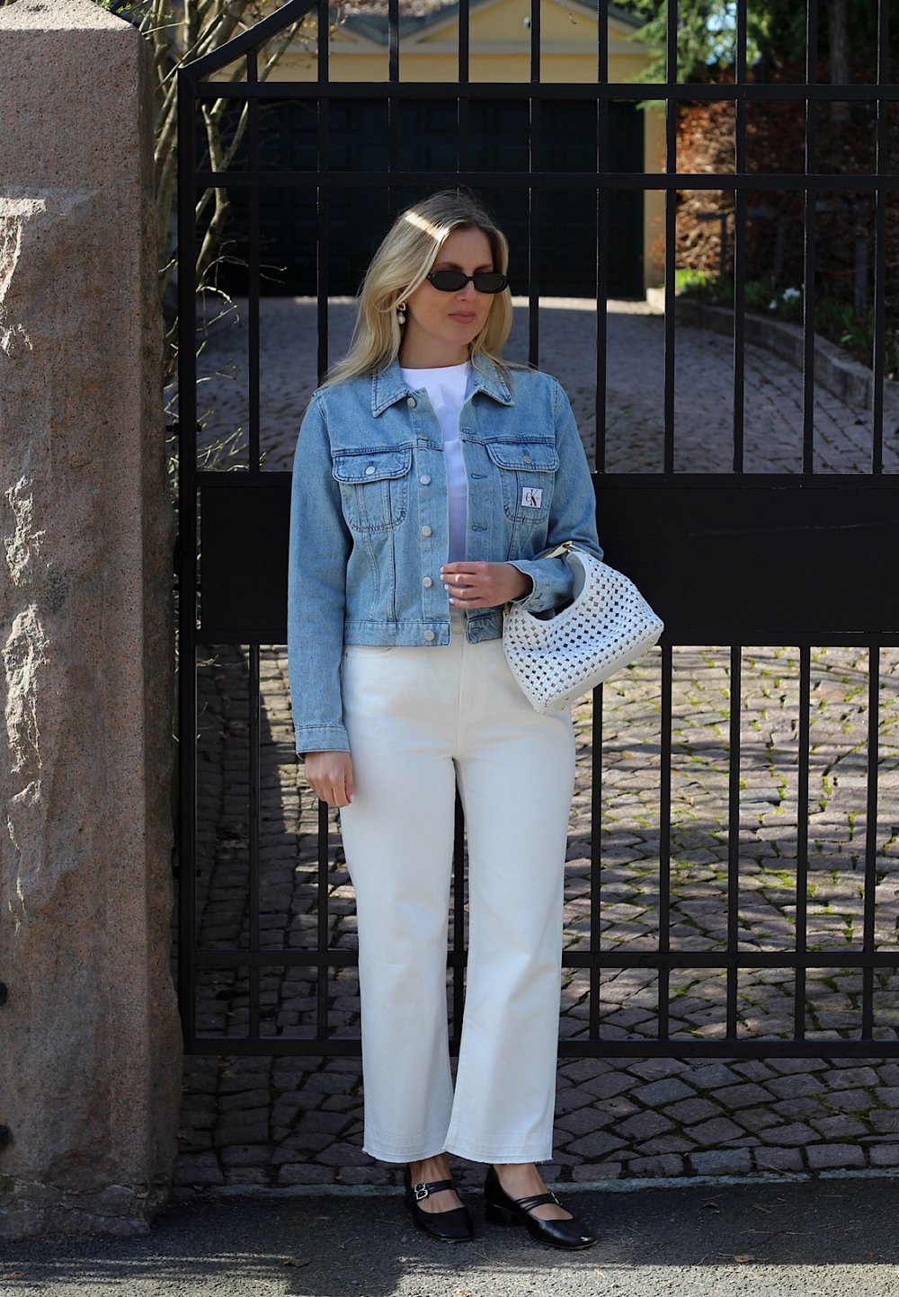 Veste en denim bleu clair, chemise blanche, pantalon large blanc, chaussures noires avec bride et sac blanc à motif perforé, devant un fond de portail.