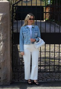 Veste en denim bleu clair, chemise blanche, pantalon large blanc, chaussures noires avec bride et sac blanc à motif perforé, devant un fond de portail.