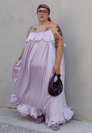 Persona con tatuajes lleva un vestido fluido color lavanda, pañuelo en la cabeza, gafas rojas, lleva un bolso oscuro y está de pie contra una pared lisa y clara.