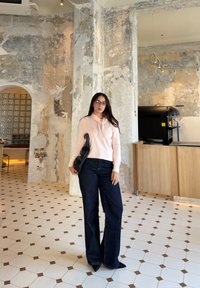 Un haut à manches longues rose clair avec un détail volanté, associé à un jean large bleu foncé et un sac à épaule noir, sur fond d'un mur texturé.