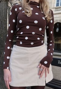 Femme portant un haut marron à manches longues avec des pois blancs et une mini-jupe beige côtelée, debout près d'un arbre et d'un banc en plein air.