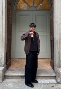 Osoba v tmavě hnědém cardiganovém svetru přes černé tričko, černých širokých kalhotách a botách, držící nápoj, před světle zelenými dveřmi.