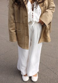 Gaišbrūna zamšāda jaka ar frizēm, balta mežģīņu blūze un brīvi piegrieztas baltas bikses. Mīkstas baltas čības papildina apģērbu.