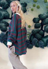Blondynka z wstążką we włosach, ubrana w zielono-czerwoną, w oversize'owym stylu sweter oraz beżowe spodnie, stojąca przed teksturowaną zieloną ścianą.