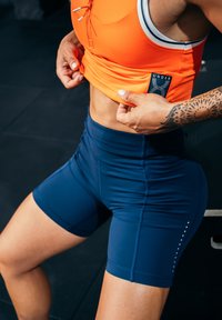 Oranžs sporta krūšturis ar priekšējo regulējamo auklu un tumši zili augsti sēdoši šorti ar sānu šuvēm un maziem punktiem.