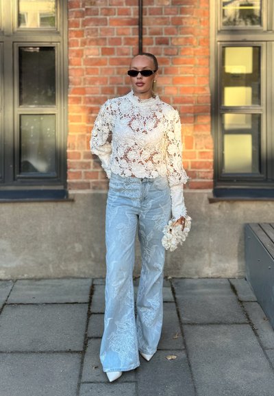Mujer con top de encaje floral blanco, vaqueros bordados de pierna ancha azul claro, zapatos blancos puntiagudos, gafas de sol negras, sosteniendo un bolso blanco con flores.