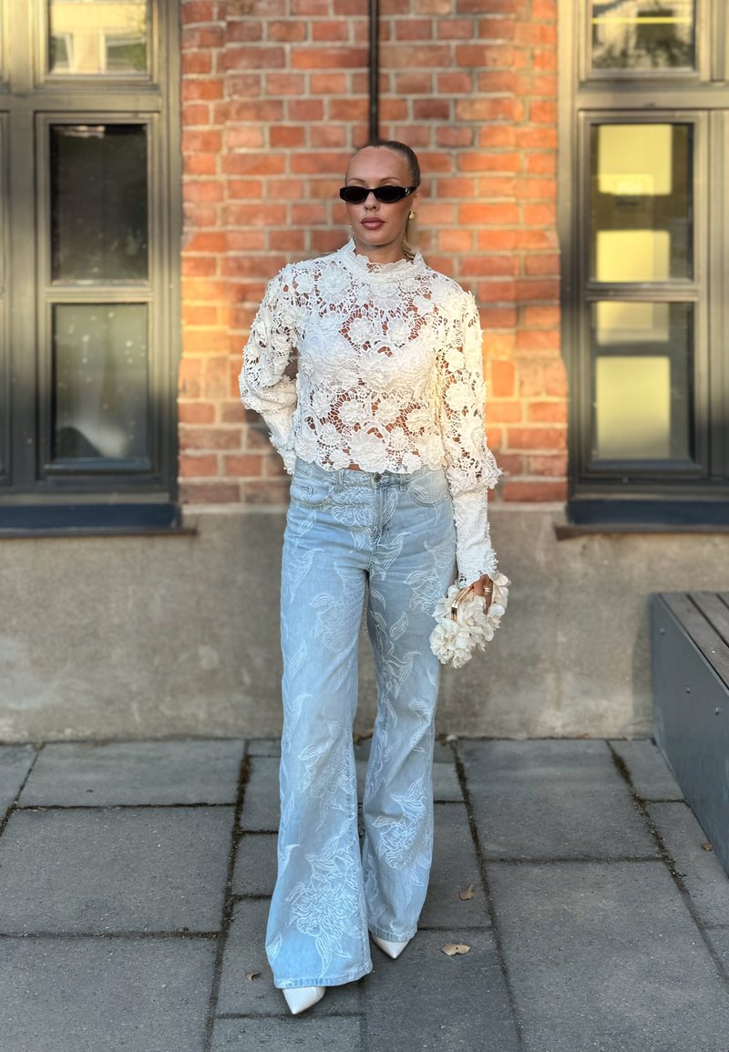 Femme portant un haut en dentelle fleurie blanche, un jean bleu clair brodé à jambes larges, des chaussures blanches pointues, des lunettes de soleil noires, tenant un sac à main blanc à fleurs.
