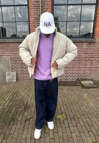 Beige pufferjack met oranje voering, paarse gebreide trui, donker denim broek en witte sneakers. Pet heeft "NA" in paars.