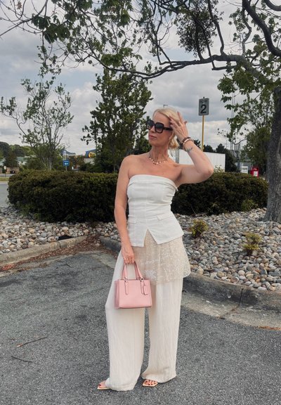 Mujer con atuendo blanco sin tirantes sosteniendo un bolso rosa, ajustándose las gafas de sol, de pie en la acera cerca de árboles y arbustos bajo un cielo nublado.