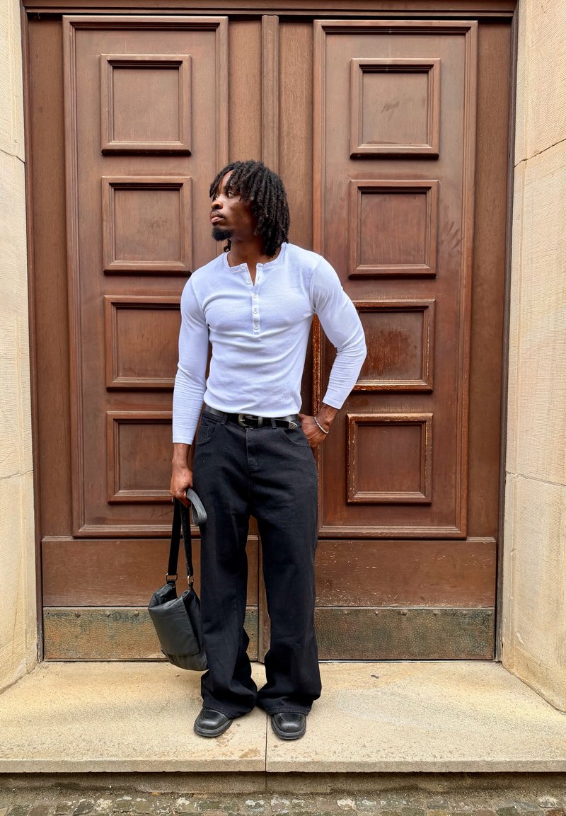 Uomo con dreadlocks che indossa una camicia bianca a maniche lunghe e pantaloni neri, in piedi con una borsa nera davanti a grandi porte doppie in legno.