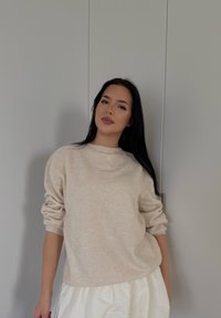 Kremno beljen pleteni pulover z dolgimi, nekoliko prevelikimi rokavi, v kombinaciji z belim, strukturiranim krilom. Nevtralen ozadje in mehka osvetlitev.