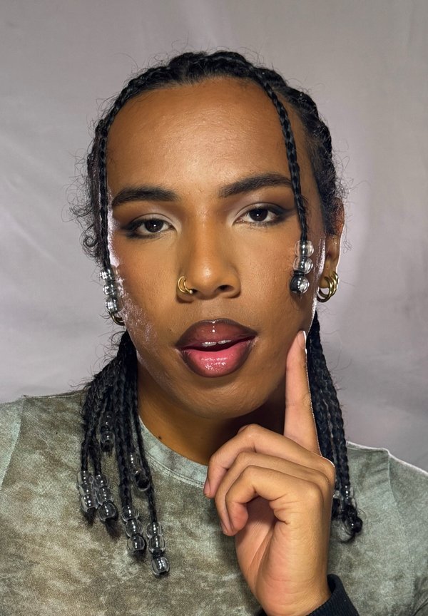 Persona con cabello trenzado adornado con cuentas transparentes, con anillo en la nariz y pendientes, tocándose la barbilla, vistiendo una blusa verde grisácea contra un fondo claro.