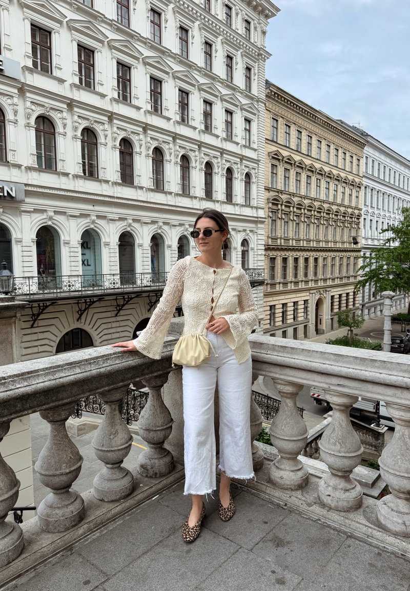 Femme en lunettes de soleil, haut en dentelle beige, pantalon blanc coupé court, chaussures à motif léopard et sac jaune, debout sur une balustrade en pierre avec des bâtiments européens ornés en arrière-plan.