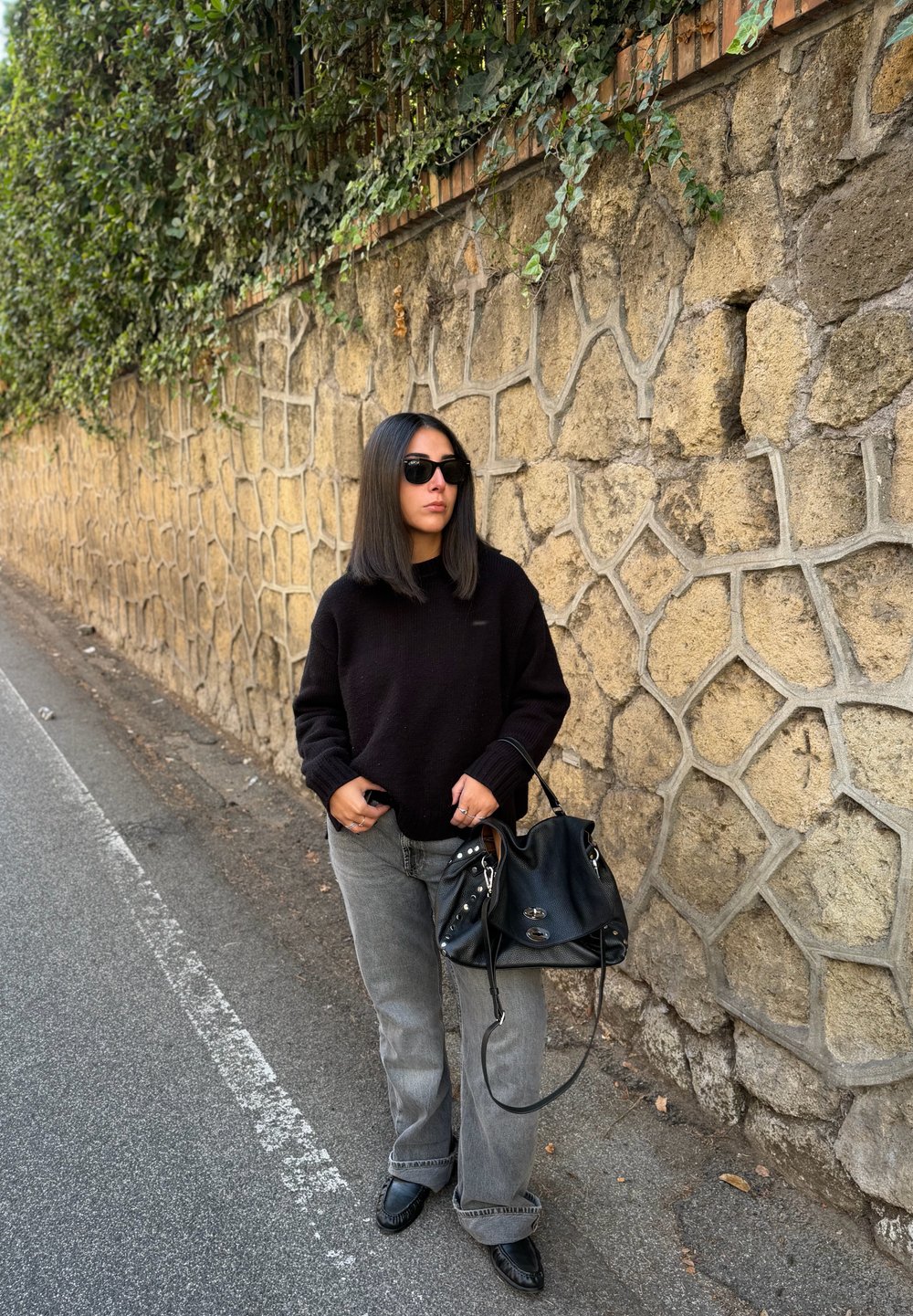 Fekete pulóver, szürke bő szárú farmernadrág, fekete kézitáska fém kiegészítőkkel és napszemüveg. Kőfal háttér zöld növényzettel.