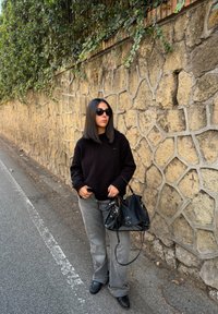 Fekete pulóver, szürke bő szárú farmernadrág, fekete kézitáska fém kiegészítőkkel és napszemüveg. Kőfal háttér zöld növényzettel.