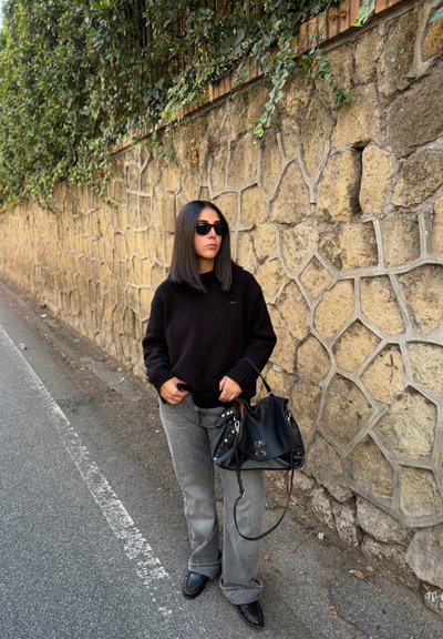 Suéter negro, jeans de pierna ancha gris, bolso negro con detalles metálicos y gafas de sol. Fondo de pared de piedra con vegetación.