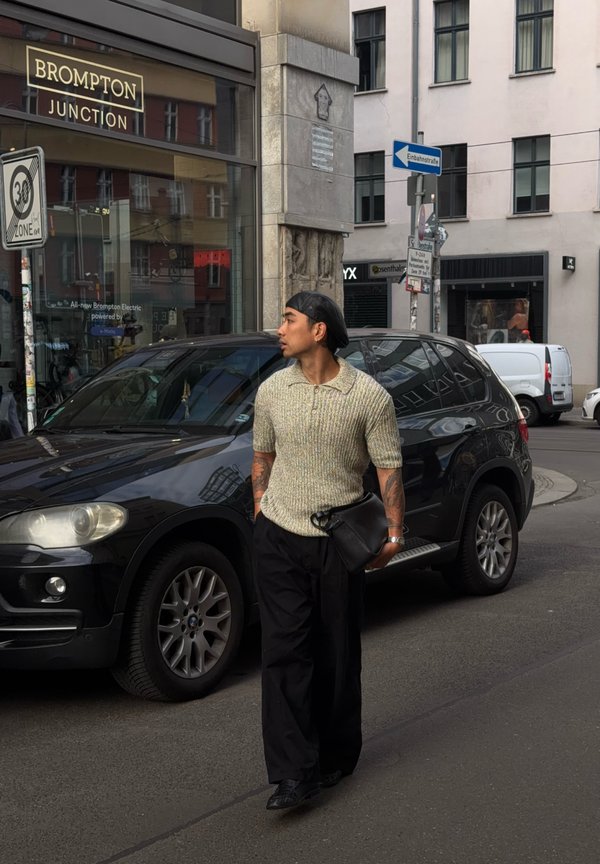 Uomo con tatuaggi che indossa un berretto, una maglia lavorata a maglia e pantaloni neri cammina per la strada della città vicino a un SUV nero parcheggiato e al negozio Brompton Junction.