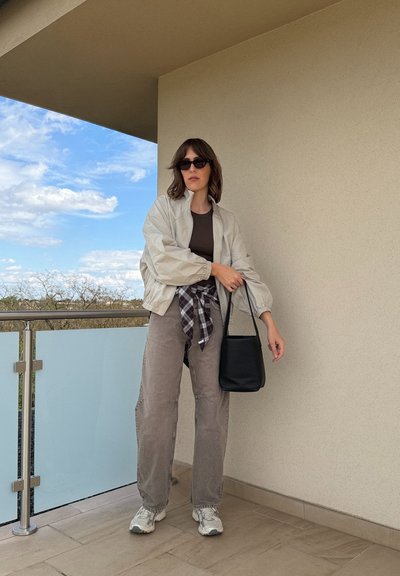 Mujer con gafas de sol, chaqueta ligera, camiseta marrón, camisa de cuadros anudada a la cintura, pantalones grises, zapatillas deportivas, sosteniendo un bolso negro en un balcón bajo una pared beige.