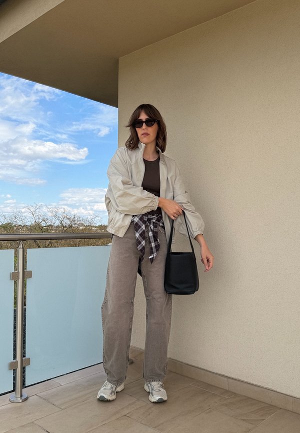 Vrouw met zonnebril, licht jasje, bruin topje, geruite blouse in de taille gestrikt, grijze broek, sneakers, draagt een zwarte handtas op een balkon onder een beige muur.