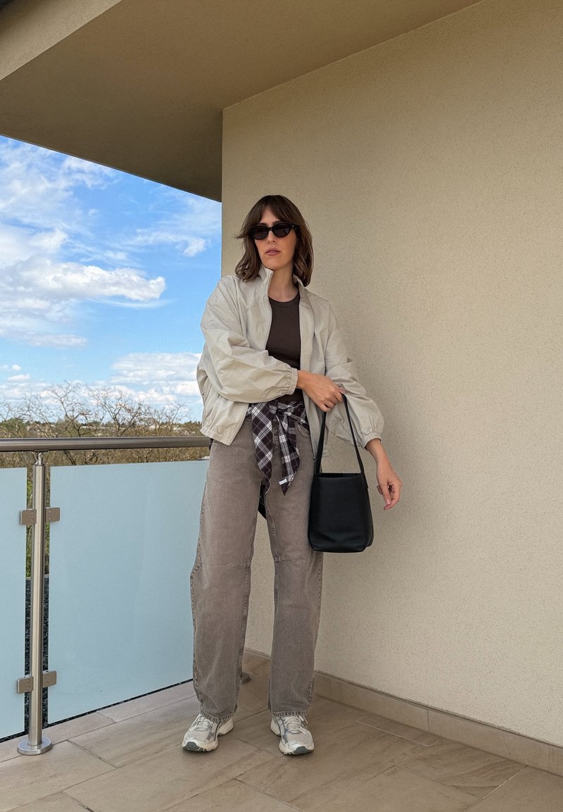 Žena sa sunčanim naočalama, laganom jaknom, smeđom majicom, kariranom košuljom vezanom u struku, sivim hlačama, tenisicama, drži crnu torbu na balkonu ispod bež zida.