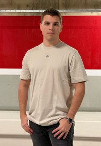 Jasnobeżowa koszulka z krótkim rękawem z małym szarym logo na piersi, zestawiona z ciemnymi dżinsami. Betonowe tło z czerwonymi akcentami.