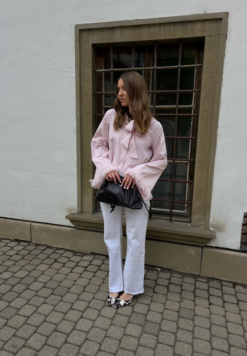 Femme aux longs cheveux bruns portant un chemisier rose à volants et un pantalon blanc, tenant un sac noir, debout dans une rue pavée près d'une fenêtre grillagée.