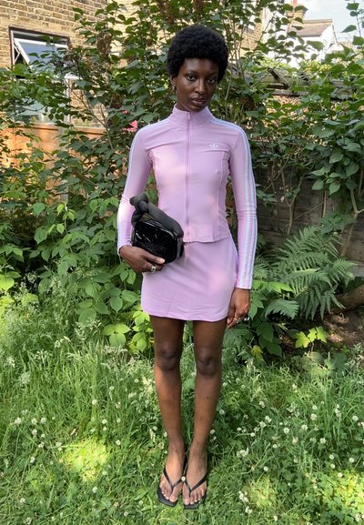 Camiseta deportiva rosa con cremallera y mangas largas, falda corta a juego y un bolso cruzado negro, todo ello en un fondo de follaje verde.