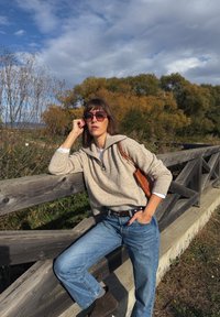 Svetlo bež pletena jopica s pol zadrgo, modri kavbojki, rjavi semiš škornji in tkan narančast nahrbtnik, postavljeni pred leseno ograjo in drevesi.