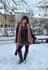 Doudoune marron avec bordure en fourrure, écharpe rose, jupe plissée grise, collants bordeaux et bottes grises montantes, dans un environnement enneigé en plein air.