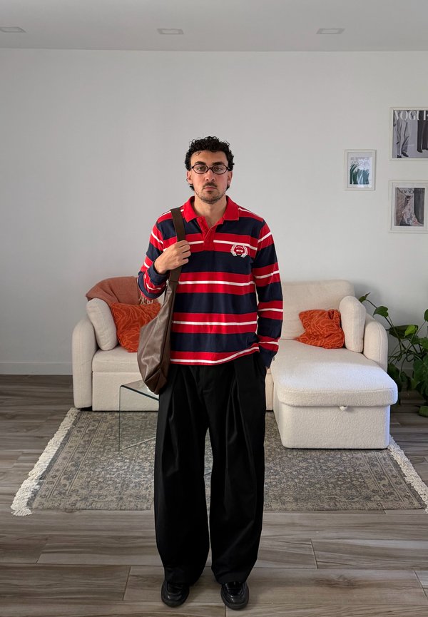 Mann mit Brille, rotem und marineblau gestreiftem Hemd, schwarzen weiten Hosen und brauner Tasche steht im Wohnzimmer mit weißem Sofa und orangen Kissen.