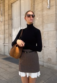 Černý rolák, kostkovaná sukně, bílý volánkový lem, hnědá taška s výřezem na uchu, sluneční brýle a kruhové náušnice, na kamenném pozadí.