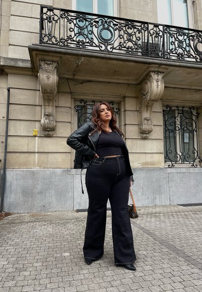 Chaqueta de cuero negra sobre un top negro corto, combinada con unos jeans de mezclilla oscuros de tiro alto y campana. Los detalles notables incluyen costuras claras y tacones puntiagudos.