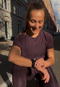 Czarny smartwatch na nadgarstku, fioletowa sportowa koszulka, teksturowana tkanina, siedzący na chodniku z promieniami słońca rzucającymi cienie, z widokiem na miasto w tle.