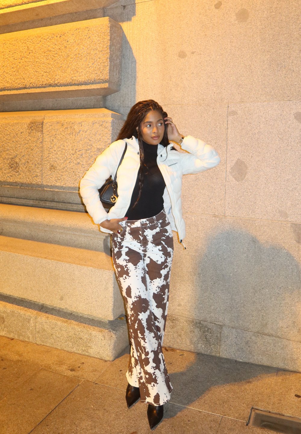 Λευκή φούτερ τζάκετ, μαύρο πουλόβερ με ψηλό λαιμό, καφέ και λευκά φλαρέ παντελόνια με σχέδιο και μαύρες γόβες. Μικρή τσάντα στον ώμο.