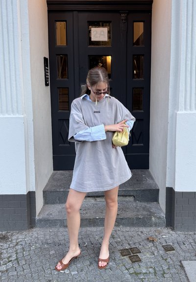 Vestido de camiseta gris de gran tamaño con un logo, superpuesto a una camisa de rayas azul claro. Sandalias de tacón marrones y un bolso amarillo completan el look.