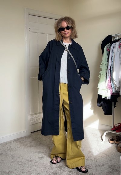 Abrigo largo azul marino con grandes botones y bolsillos laterales, combinado con pantalones mostaza de pierna ancha y sandalias negras. Diseño minimalista.
