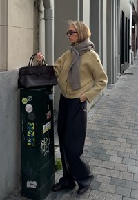 Oversized béžový žebrovaný kardigan, šedý šátek, tmavé široké kalhoty, černá kabelka, stojící vedle zelené utility krabice v městském prostředí.