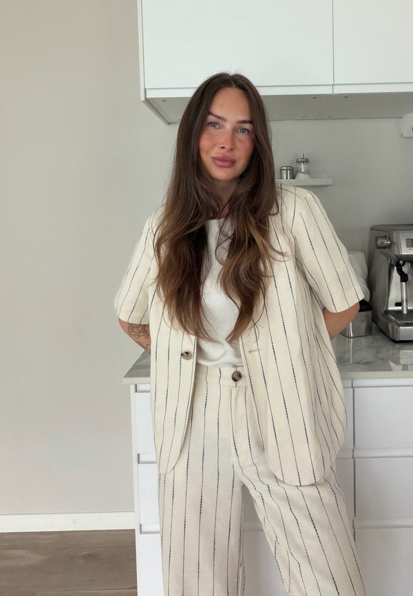 Mujer con cabello largo y castaño que lleva un traje blanco de rayas, de pie en una cocina moderna con máquina de café y armarios blancos.