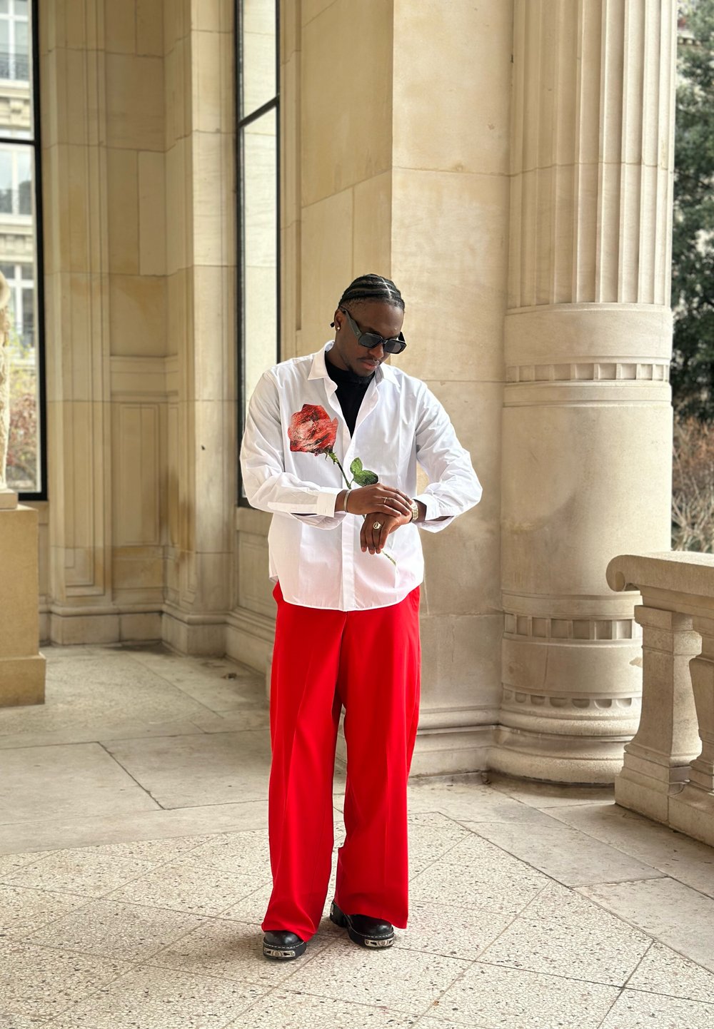 Chemise blanche boutonnée avec illustration de rose rouge, associée à un pantalon large rouge et des chaussures noires. Lunettes de soleil et montre portées.